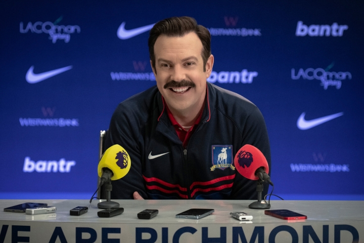 Ted Lasso senta-se em uma mesa de coletiva de imprensa na 3ª temporada.