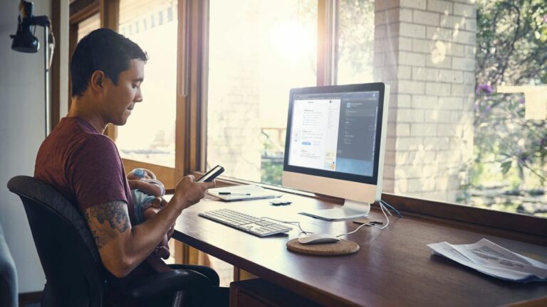 A person holds their phone while they sit at a desk with a desktop computer on top of it.