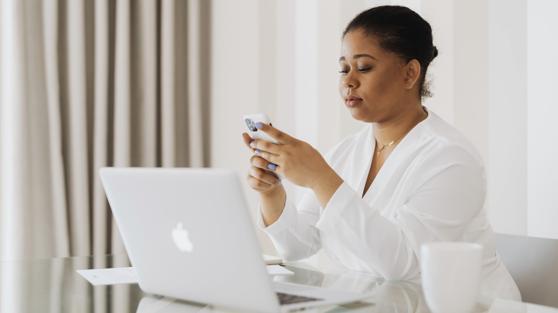 Uma mulher segura um iPhone enquanto usa um MacBook em uma mesa.