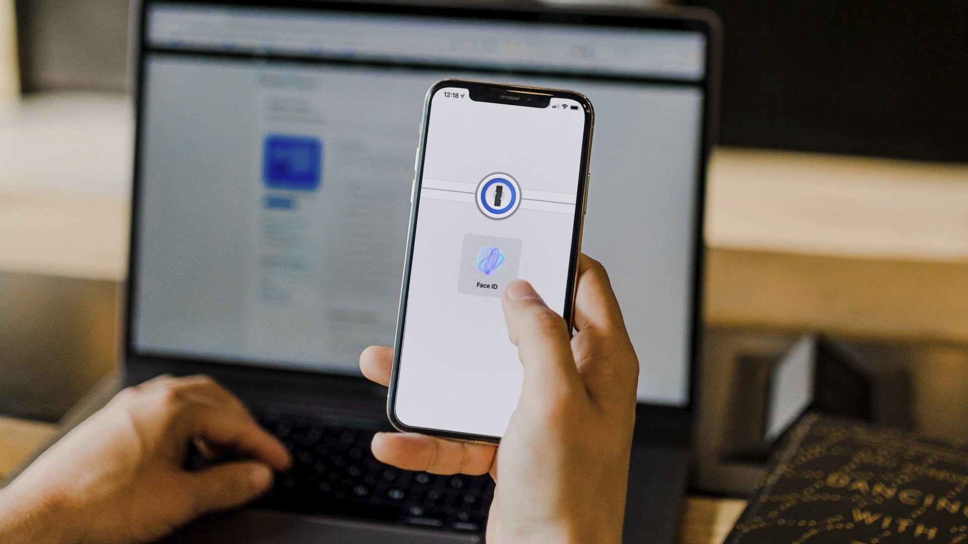 Uma pessoa segura seu iPhone e desbloqueia sua conta 1Password usando o Face ID.  Ao fundo, um notebook.