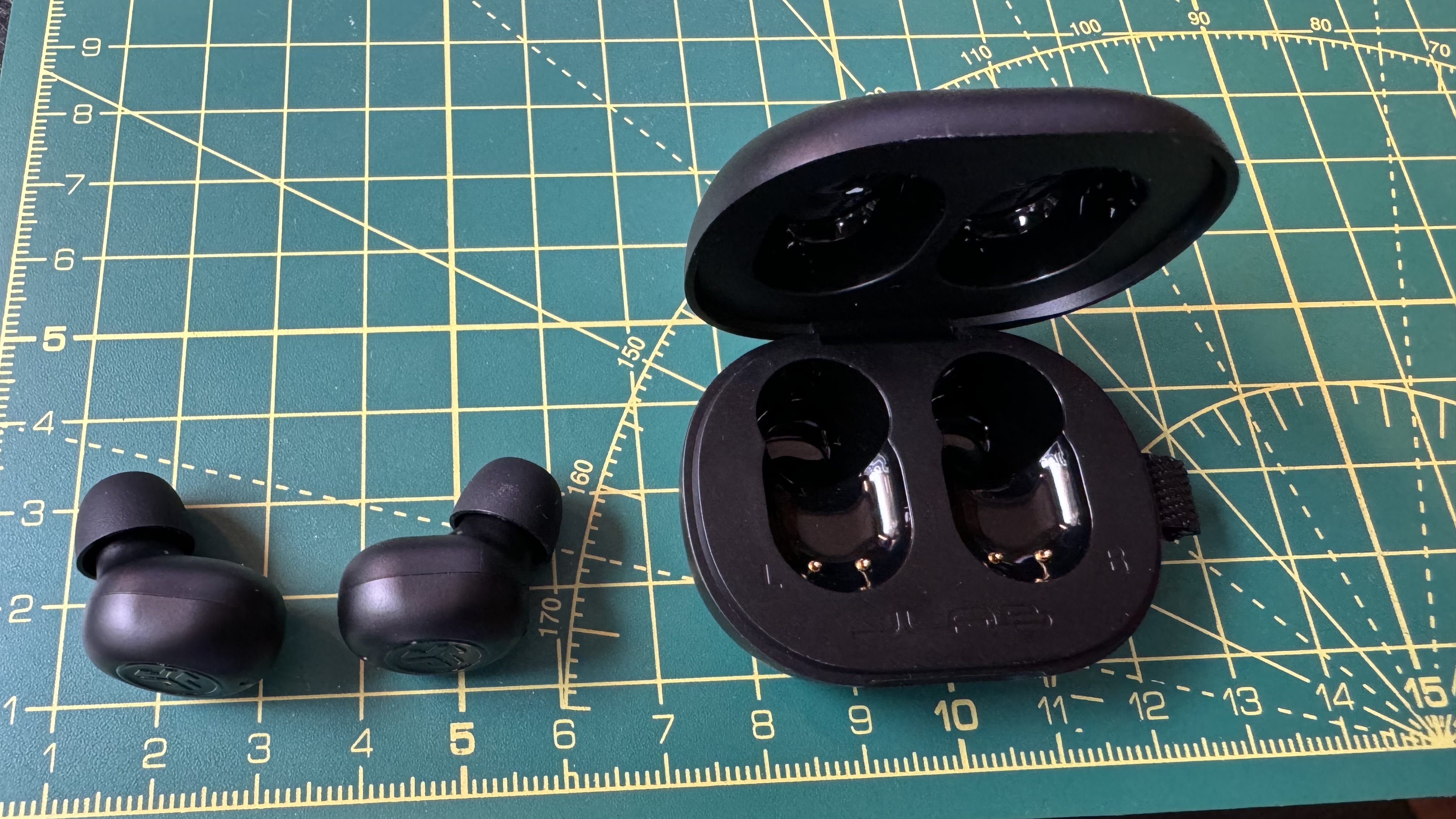 Fones de ouvido sem fio verdadeiros JLab JBuds Mini em um livro e tapete técnico, com mão e AirPods para comparação.