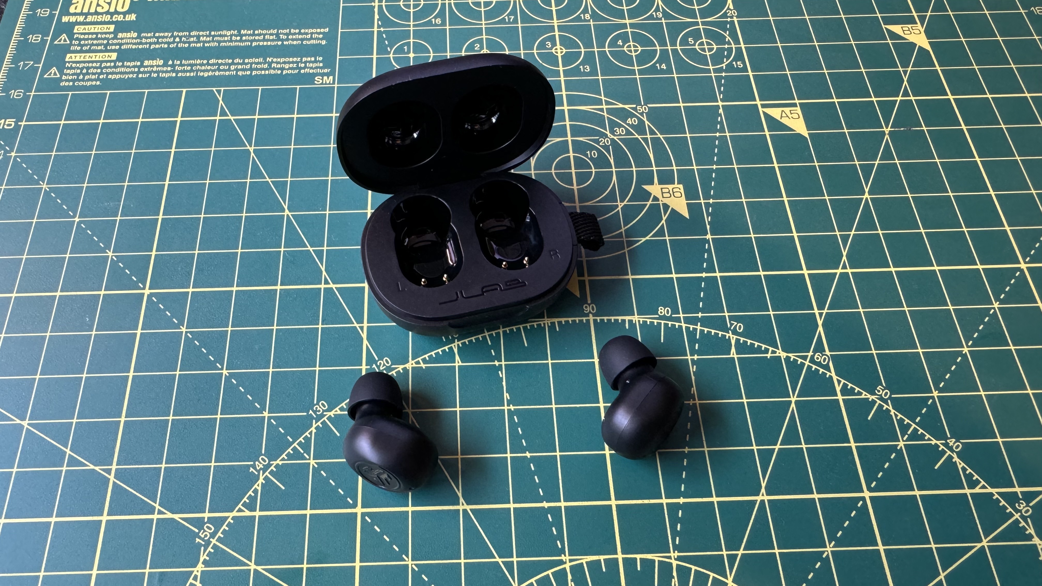 Fones de ouvido sem fio verdadeiros JLab JBuds Mini em um livro e tapete técnico, com mão e AirPods para comparação.