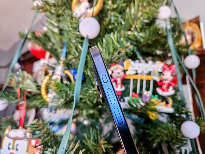 iPhone 15 Pro em Blue Titanium mostrando vista lateral dos botões de ação e volume em frente a uma árvore de Natal.