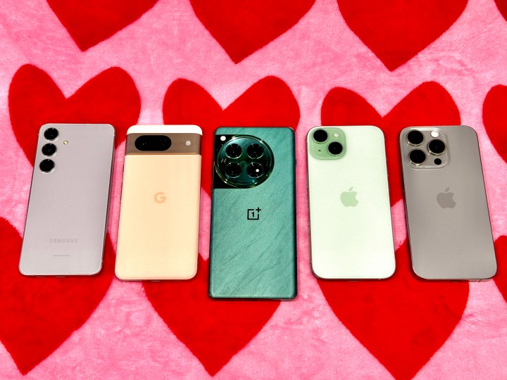 Samsung Galaxy S24 cinza (esquerda), Google Pixel 8 em ouro rosa, Flowy Emerald OnePlus 12, iPhone 15 verde, iPhone 15 Pro cinza titânio em um cobertor de coração rosa e vermelho.